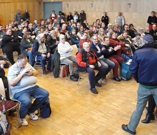 Kurz vor der Begrüßung. Die Teilnehmenden sitzen auf Stühlen oder lehnen an der Wand im Großen Saal des Freizeitheims Vahrenwald. Kurz vor der Begrüßung. Die Teilnehmenden sitzen auf Stühlen oder lehnen an der Wand im Großen Saal des Freizeitheims Vahrenwald.